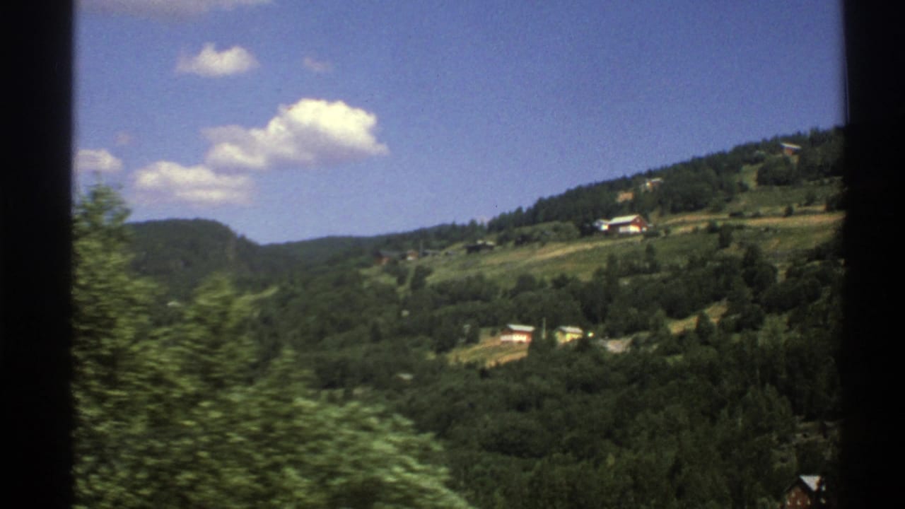 1976 8mm home-movie view of green Norwegian hills, pine forest, distant houses, and passing roadside foliage beneath scattered clouds.