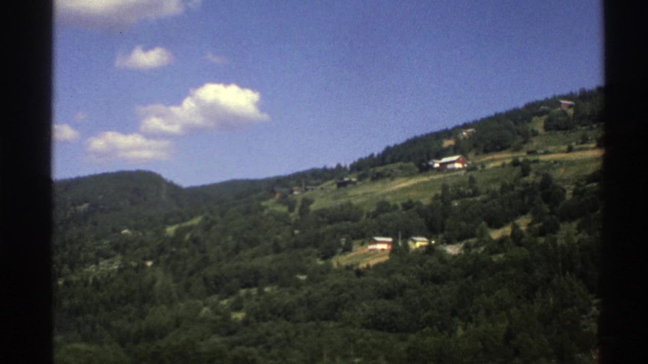 Wide view of green valley slopes, tree cover, and small houses beneath scattered clouds.