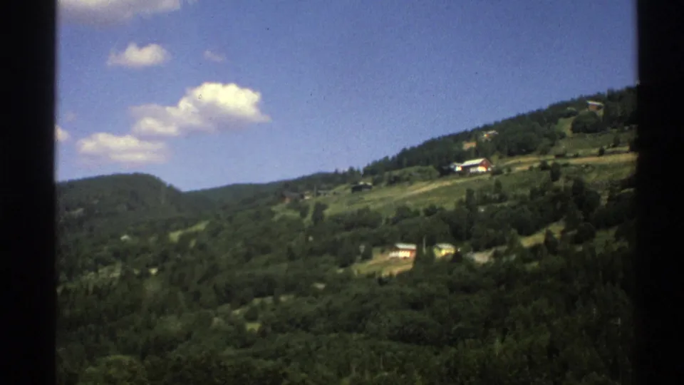 Norwegian rural hillside in 1976, with wooded slopes, small houses, green fields, and visible 8mm gate edges from a moving vehicle shot.