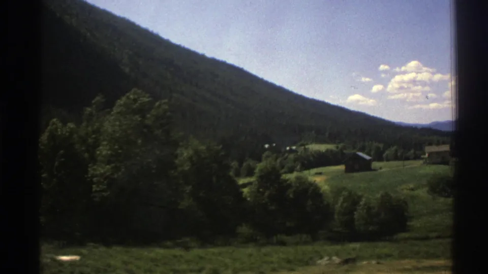 1976 8mm Norway travel view with a shaded forested mountain slope, green meadow, distant small buildings, and bright clouds overhead.