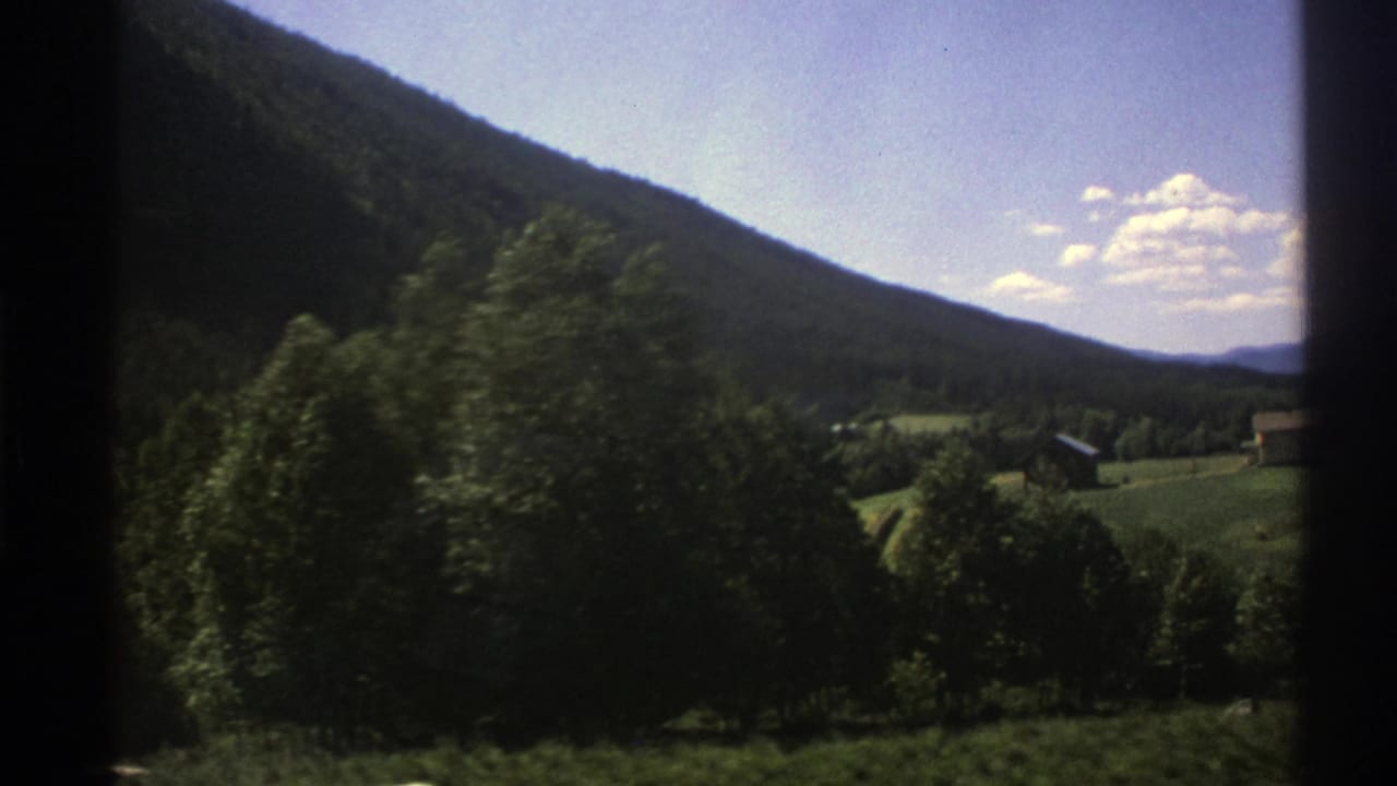 1976 Norway home-movie view of a wooded hillside and green valley with small houses, filmed from a moving vehicle.