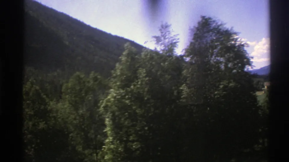 Passenger-view 8mm frame from Norway in 1976, with blurred trees, a wooded slope, and distant valley scenery.