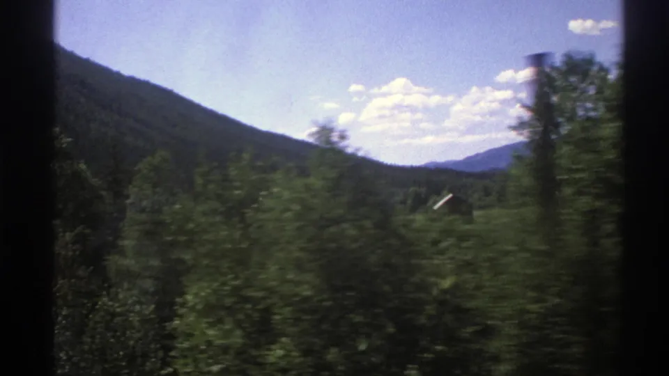 1976 Norway 8mm countryside frame with blurred trees, a sloping wooded hill, distant mountains, and scattered clouds.
