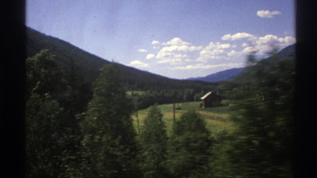 1976 Norway 8mm travel view of fields, scattered trees, small rural buildings, and mountain slopes under a bright blue sky.