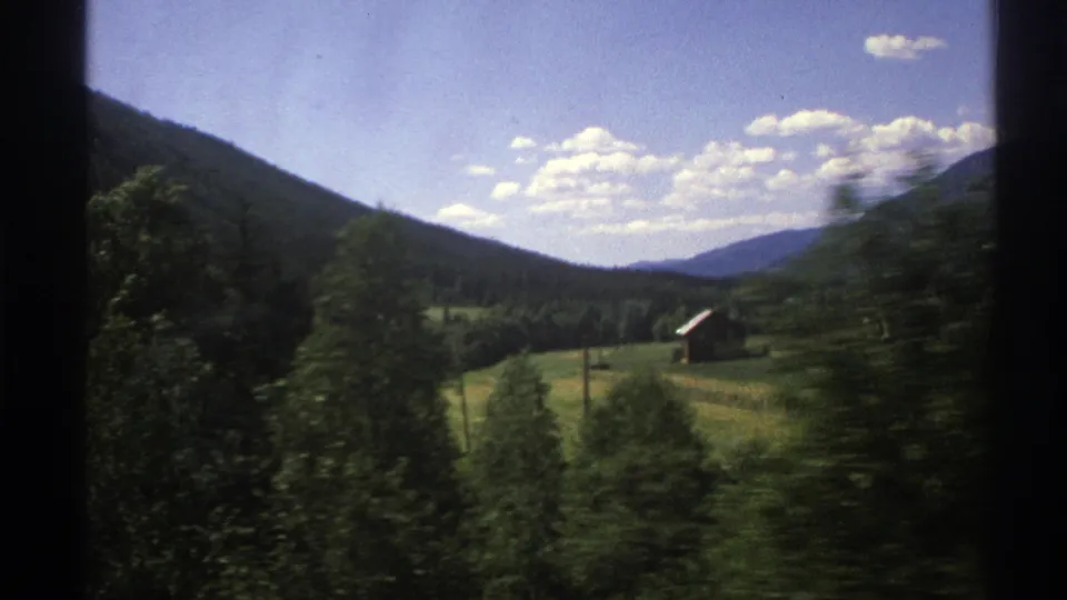 1976 Norway 8mm travel view of fields, scattered trees, small rural buildings, and mountain slopes under a bright blue sky.