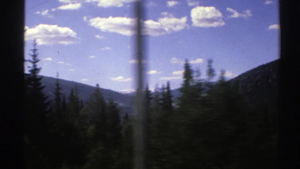 1976 Norway travel frame with blurred roadside trees, forested hills, and mountain forms beneath scattered white clouds.