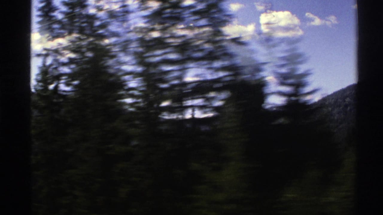 1976 Norway 8mm frame showing dark conifers blurred by vehicle motion with a mountain slope beyond and bright clouds overhead.