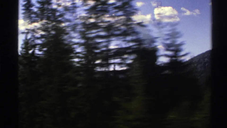 1976 Norway 8mm frame showing dark conifers blurred by vehicle motion with a mountain slope beyond and bright clouds overhead.