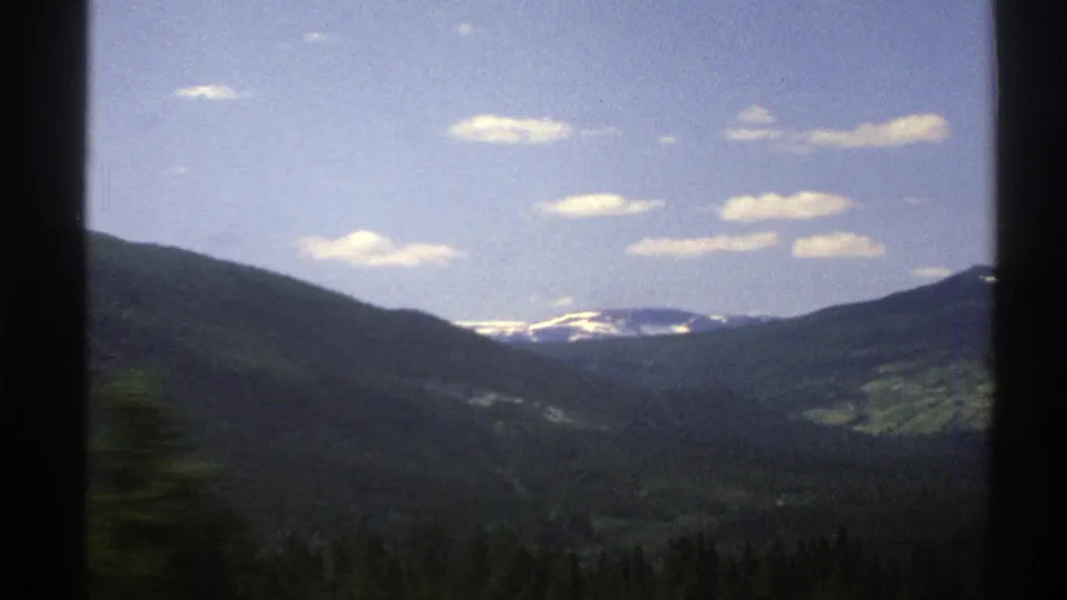 Scenic Norwegian mountain valley from a moving vehicle, with distant snow patches and scattered clouds visible through the window.