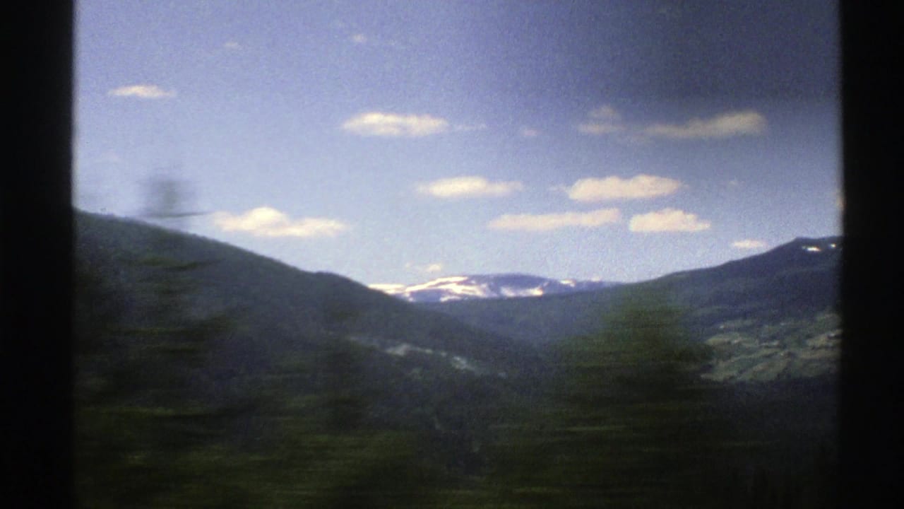 Moving valley view with blurred roadside trees, distant mountains, snow patches, and blue sky.