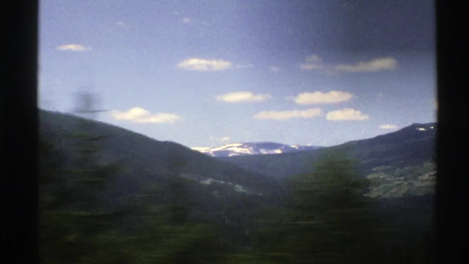 1976 Norway 8mm travel frame showing a moving car view across a green valley toward distant snow-capped mountains under a bright sky.