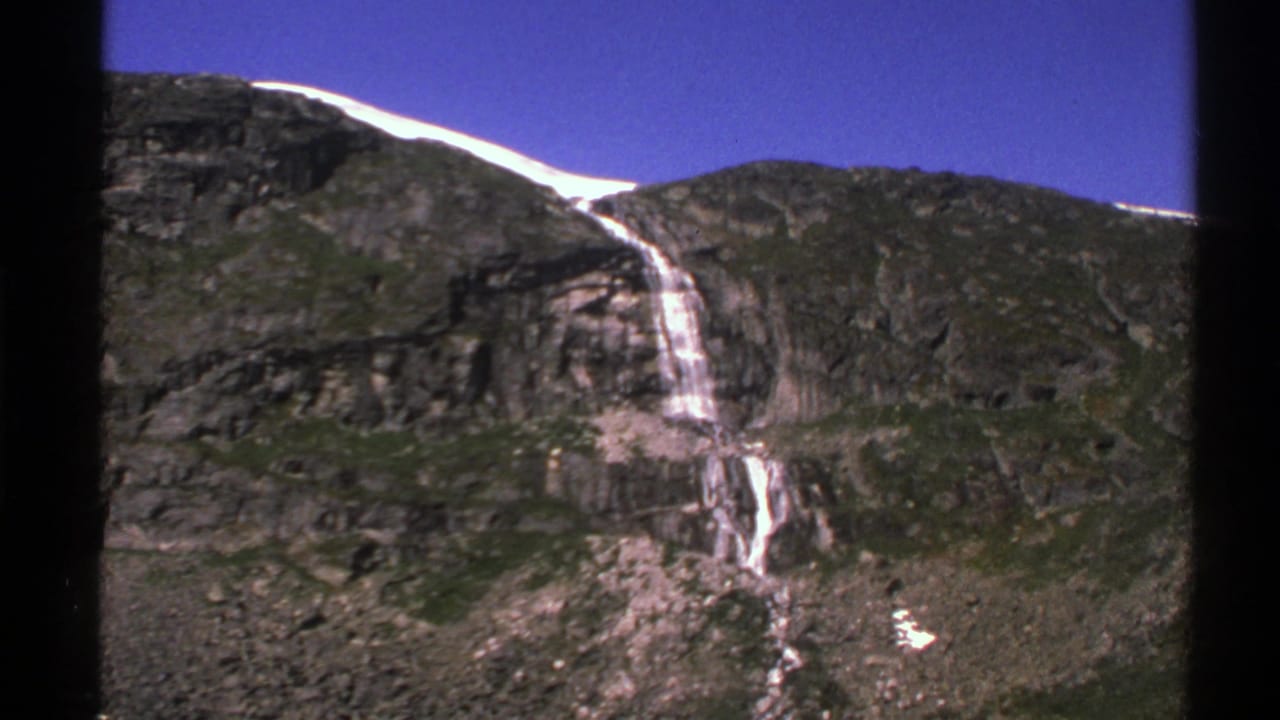 Rocky mountain face with a narrow waterfall, snow along the ridge, and deep blue sky.