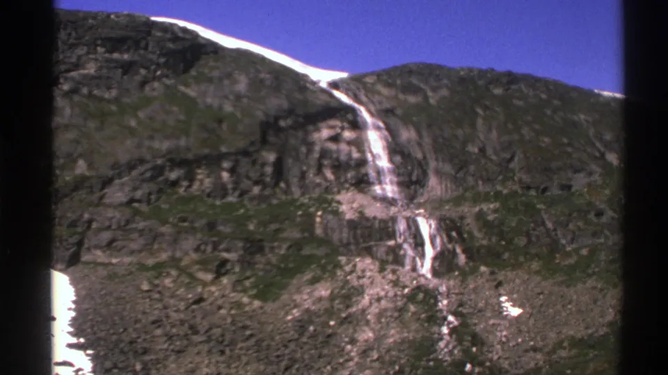 1976 Norway home-movie frame showing a closer handheld view of a waterfall running down a steep rock face with snow patches nearby.