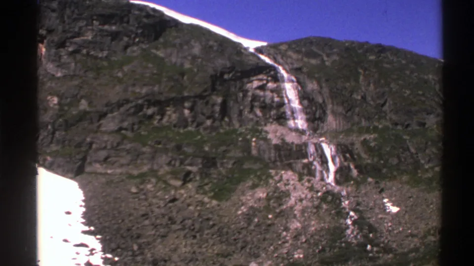 1976 Norway 8mm travel frame of a mountain waterfall crossing dark rock and green slopes, with snow patches and film gate borders visible.