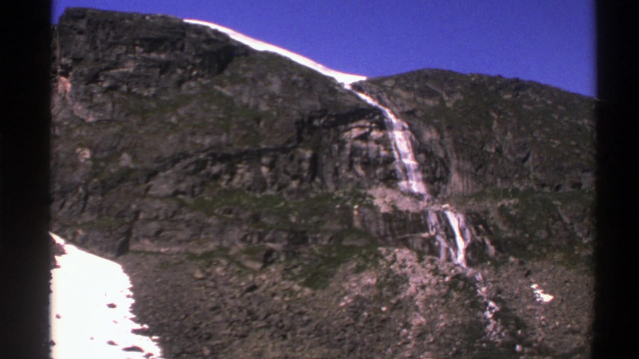 Rocky mountain cliff with waterfall, snow patches, and blue sky.