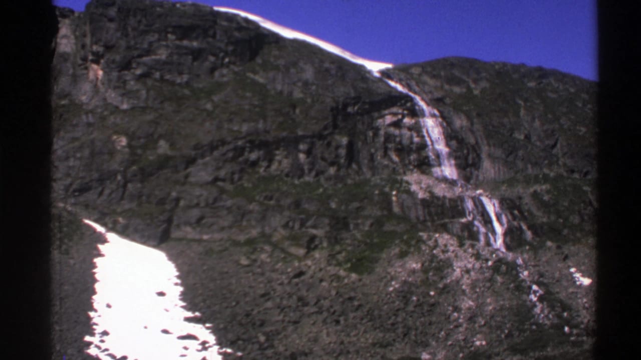 Mountain slope with waterfall, dark rocks, snow, and clear blue sky.