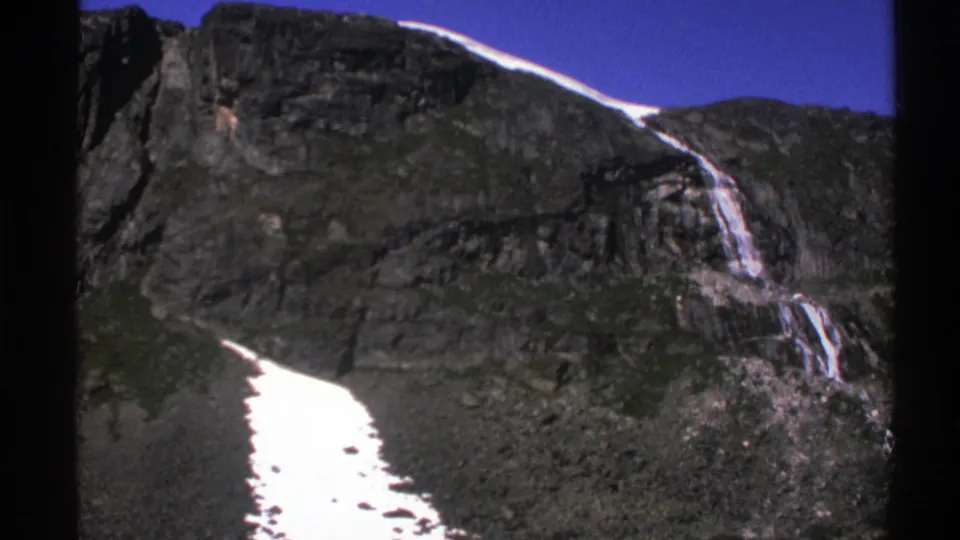1976 Norway 8mm frame showing a closer view of dark alpine rock, a descending waterfall, and a bright snowfield in the foreground.