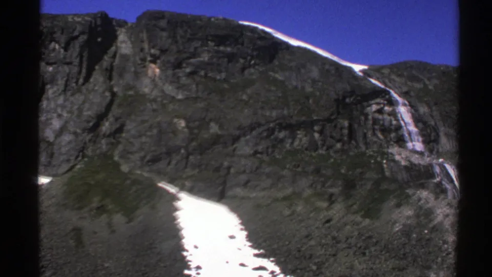 1976 Norway 8mm frame showing a rocky mountaintop slope, waterfall, snow patch, and saturated blue sky.