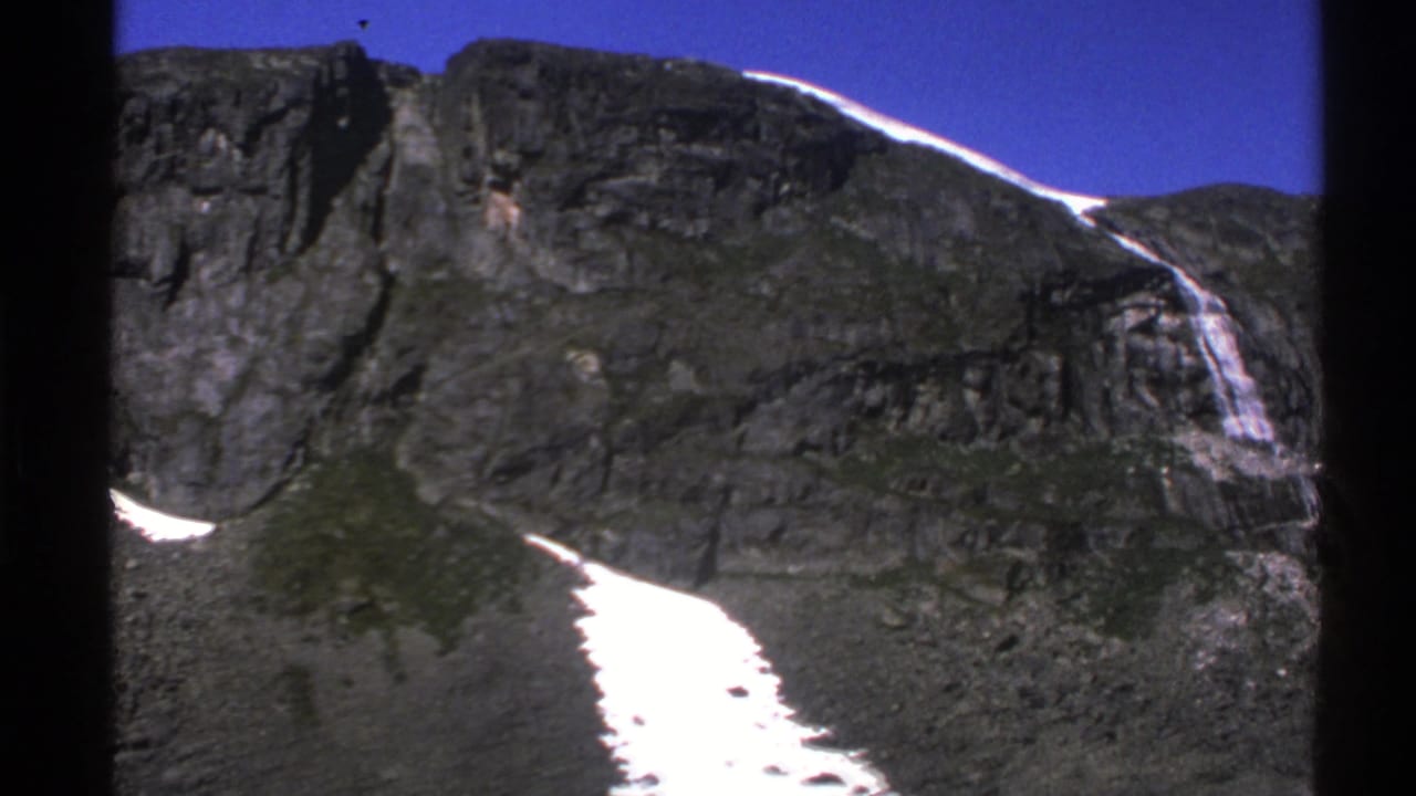 Rocky mountain slope with snow patches and a thin waterfall under blue sky.