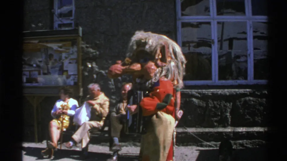 Norway 1976 frame showing a furry-costumed performer with accordion entertaining seated adult spectators outside a stone building.