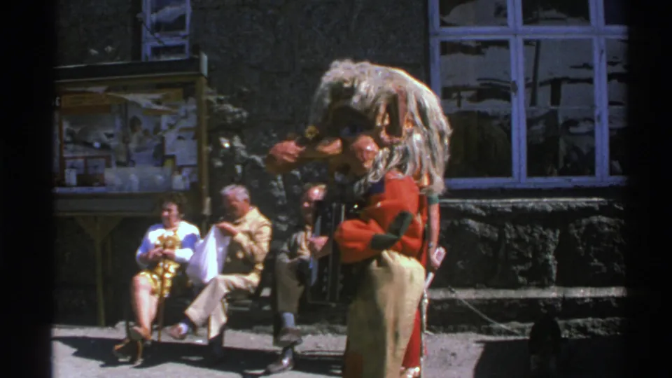 Norway 1976 home-movie frame of older spectators seated near a stone wall as a costumed accordion performer stands nearby.