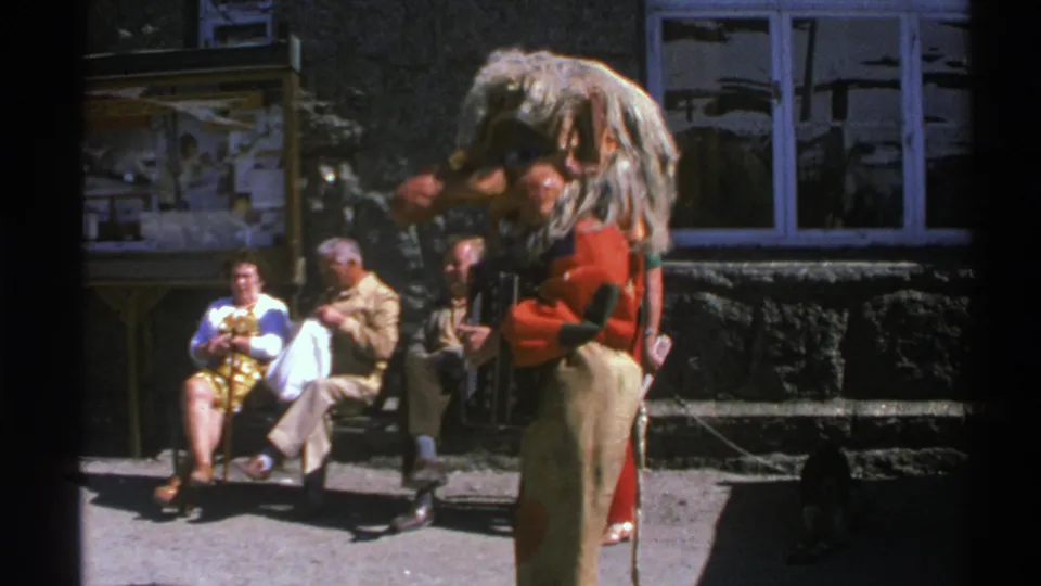 Norway 1976 frame showing a red-jacketed performer in a large furry headpiece near seated spectators outside a stone building.