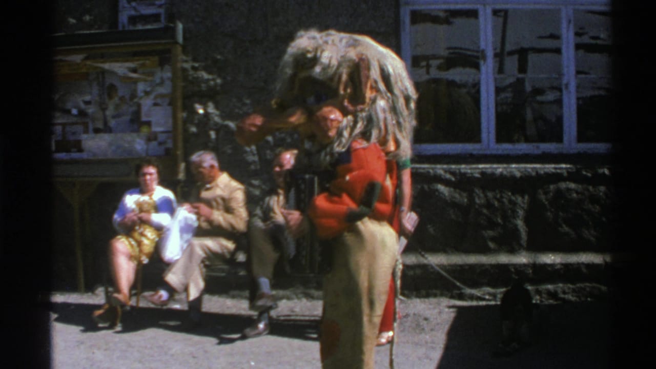 People seated and standing outside a building in bright sunlight, with a large costumed or shaggy figure in the foreground.