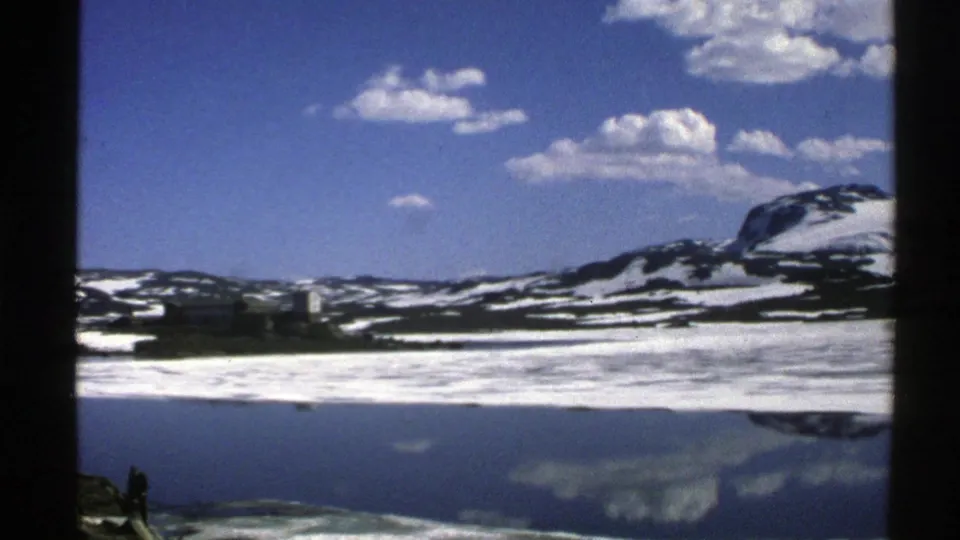 1976 Norway landscape frame with snow-dusted mountains reflected in a still lake beneath bright blue sky and small clouds.