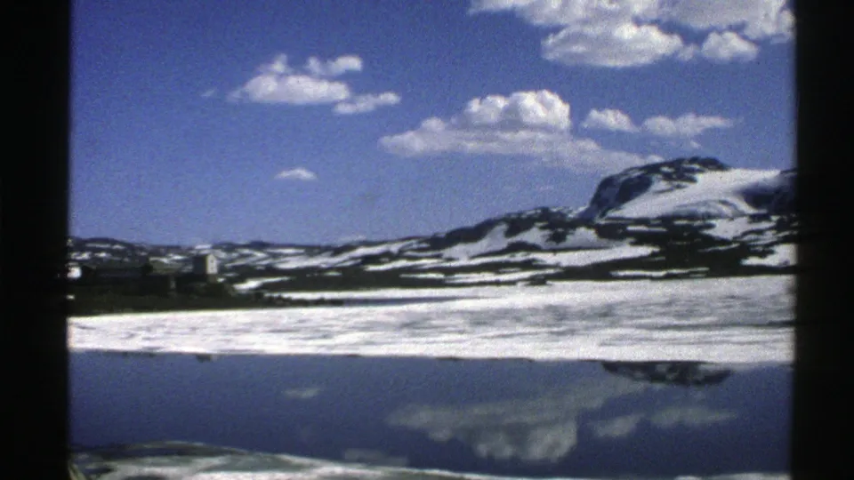 1976 Norway 8mm frame of a broad snow-covered mountain slope mirrored in a quiet lake under scattered white clouds.