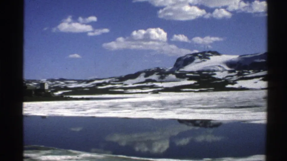 1976 Norway mountain-lake view with snow patches, cloud reflections, and a lone small figure near the icy shoreline.