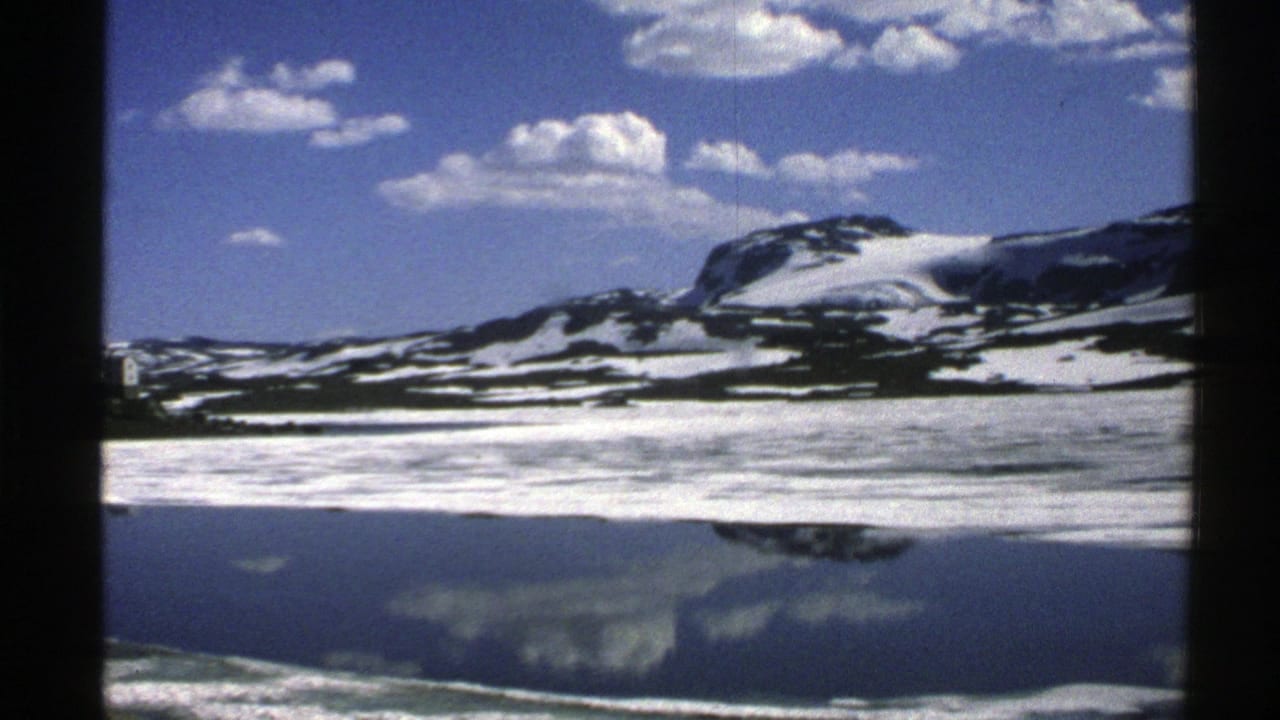 Snow-streaked mountain above a calm reflective lake under a blue sky with scattered clouds.