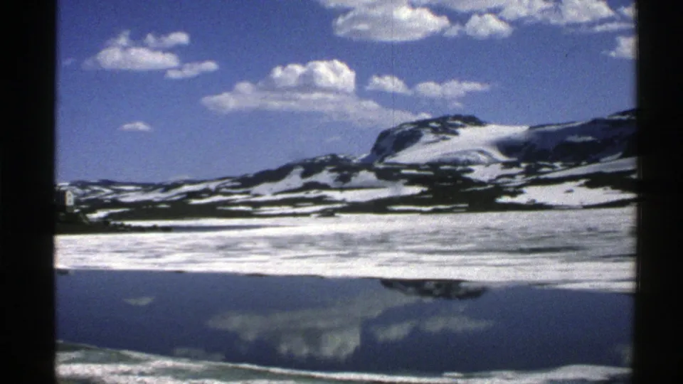 1976 Norway 8mm frame showing a snow-streaked mountain ridge reflected in a still lake beneath a bright clouded sky.