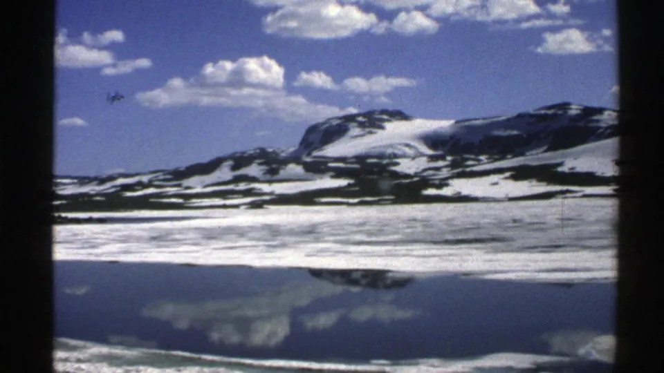 1976 Norway landscape frame with a snow-patched mountain and scattered clouds reflected across a still lake surface.