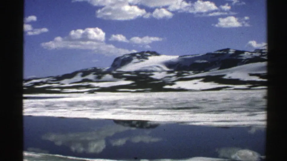 1976 Norway 8mm still with snow on mountain slopes, bright clouds overhead, and a lake reflection in the foreground.