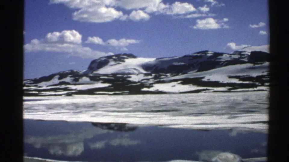 1976 Norway home-movie frame showing a broad snow-streaked mountain ridge and its reflection across a calm lake.