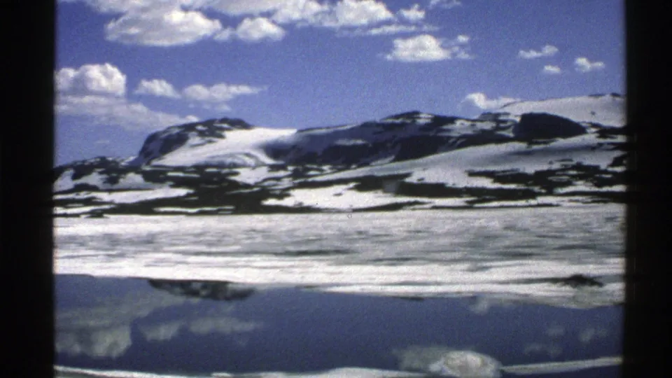 1976 Norway home-movie frame with snow-patched mountains, bright clouds, and their reflection on a quiet water surface.