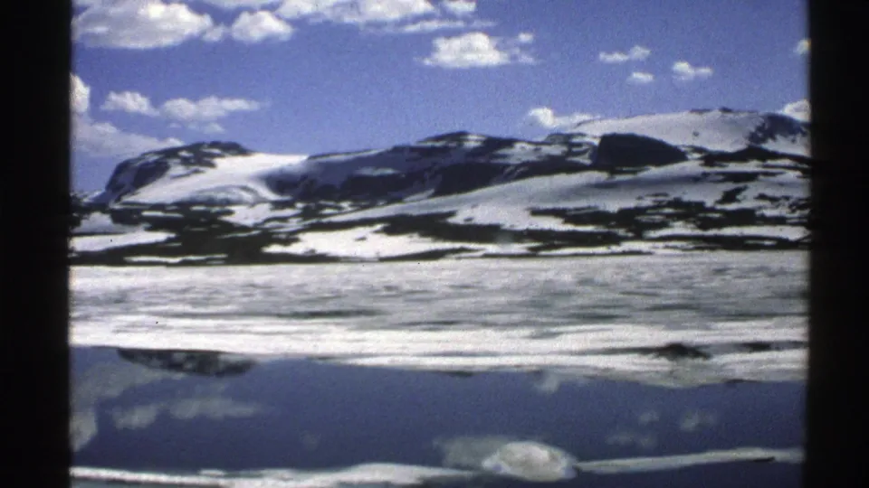 1976 Norway 8mm frame presenting a wide snow-marked mountain view with cloud reflections across calm water.