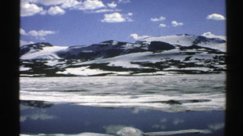 1976 Norway home-movie frame showing a snowy ridge, dark land patches, and a subdued reflection on still water.