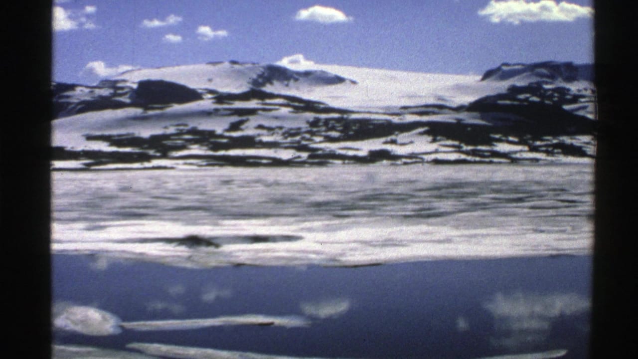 1976 Norway home-movie frame showing snow-covered mountains above a reflective frozen expanse under a blue, cloud-dotted sky.