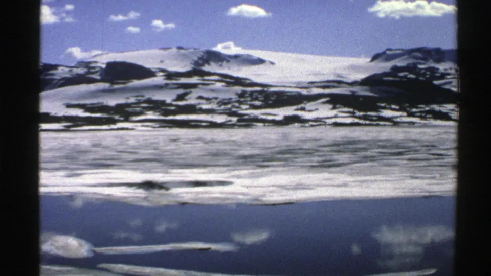 1976 Norway home-movie frame showing snow-covered mountains above a reflective frozen expanse under a blue, cloud-dotted sky.
