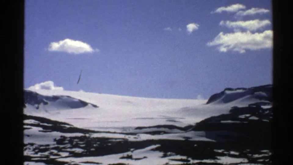 1976 Norway 8mm frame with a wide snowfield stretching between dark rocky slopes and small clouds in a clear blue sky.