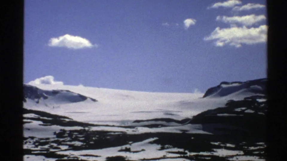 1976 Norway home-movie frame showing a snowy mountain basin, exposed rock patches, distant ridges, and scattered clouds overhead.