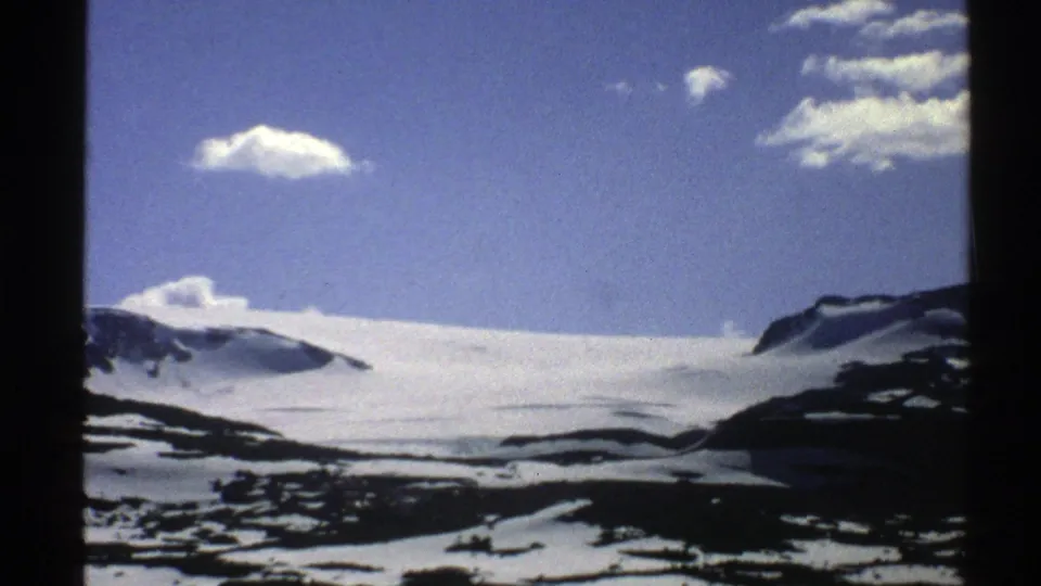 1976 Norway 8mm frame of snow-covered slopes and dark rocky ground beneath a bright blue sky with scattered white clouds.