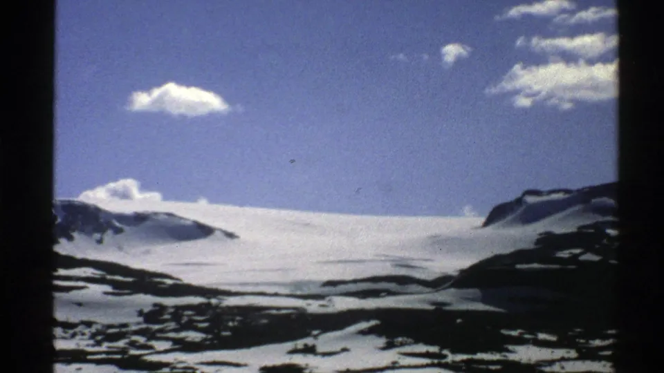 1976 Norway 8mm frame showing a bright snowfield, exposed rock, and a few clouds in a clear mountain sky.
