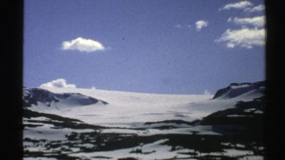 1976 Norway 8mm frame of partly snow-covered alpine terrain, dark slopes, and white clouds against a blue sky.