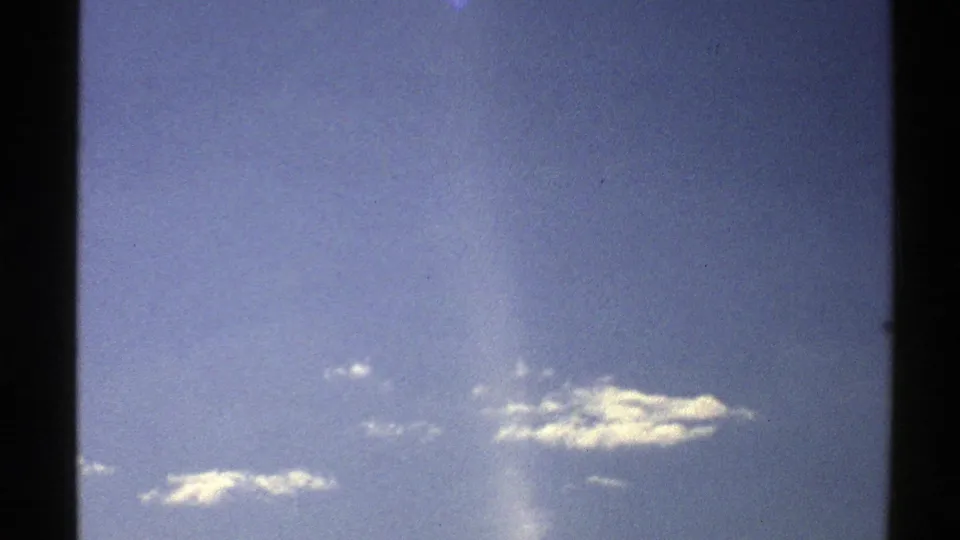 1976 Norway 8mm frame focused on open blue sky, small white clouds, visible grain, frame edge, and a vertical projection-like light flare.