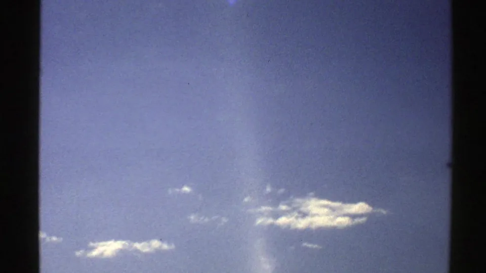 1976 Norway 8mm frame showing a broad blue sky with small cloud clusters, soft grain, slight fading, and a pale vertical flare near center.