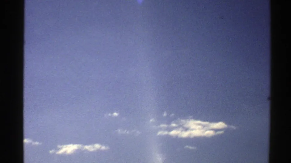 1976 Norway 8mm frame showing scattered white clouds in a blue sky, a pale vertical flare, visible grain, and dark film border along the edge.