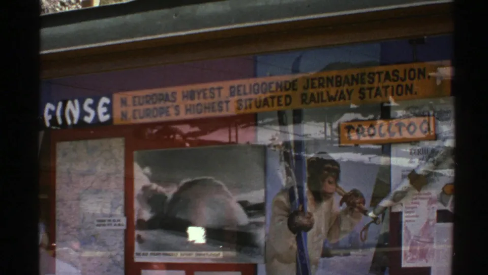 1976 Norway shop-window view with a bilingual railway-station sign, travel notices, reflections, and an illustrated monkey poster.