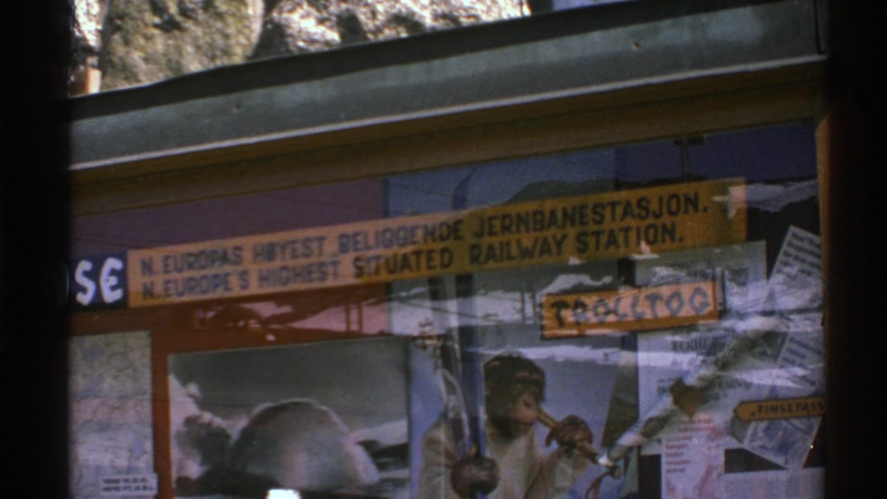 1976 Norway 8mm frame showing a bilingual railway-station sign through glass, with reflections and a chimpanzee poster beneath it.